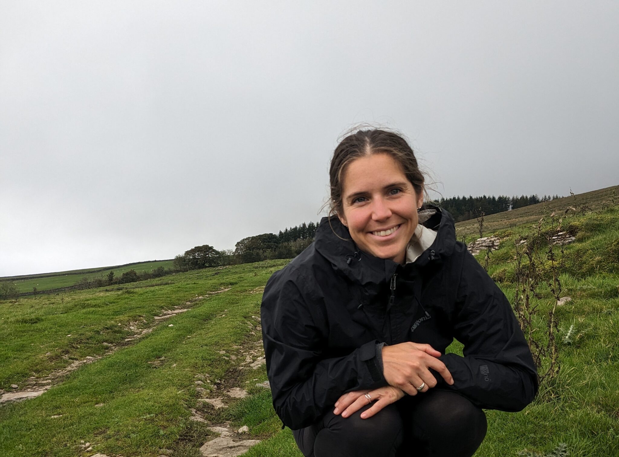 brown haired girl smiling at the camera with a black rain jacket on a green field